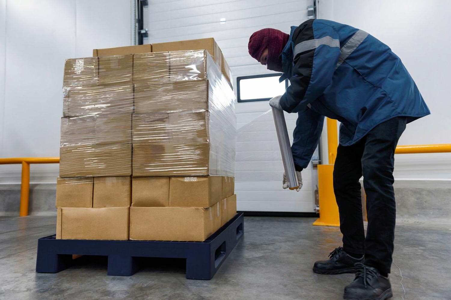 Pallet Shrink Wrapping in Pharmaceutical Warehouse Warehouse worker wrapping stacked cardboard boxes on a pallet with protective stretch film.